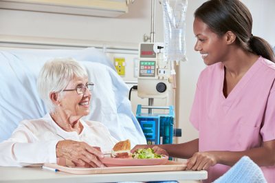 Nurse Serving Senior Female Patient Meal In Hospital Bed Smiling
